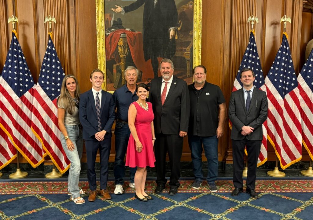 Seven people stand in a formal room with wooden walls, American flags, and a large historical portrait behind them. Four men wear suits, two men dress casually, and one woman wears a pink dress, while another wears a blouse and jeans.