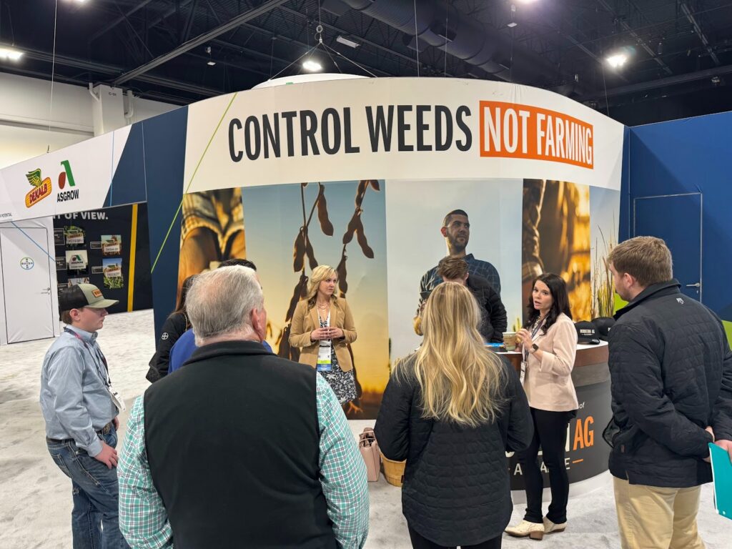 A group of people gathers at a trade show booth with a large sign reading CONTROL WEEDS NOT FARMING. The booth features agricultural branding and banners, and attendees are engaged in conversation.