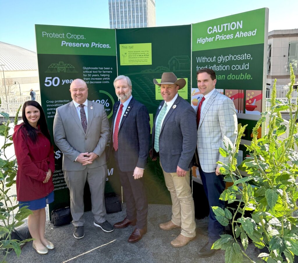 Five people stand smiling in front of a green display about crop protection and food prices, with tall plants in the foreground and buildings in the background. Four men wear suits; one woman wears a red coat.