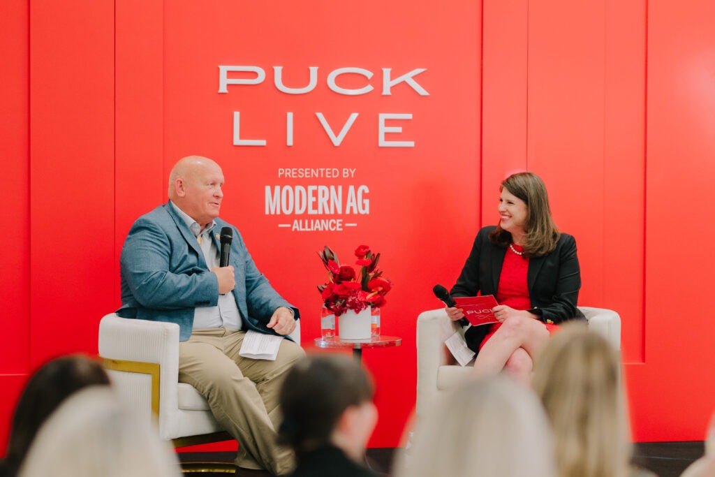 Two people sit on white chairs, holding microphones, engaged in conversation on stage against a bright red backdrop with PUCK LIVE Presented by Modern Ag Alliance written on it. A small table with red flowers is between them.