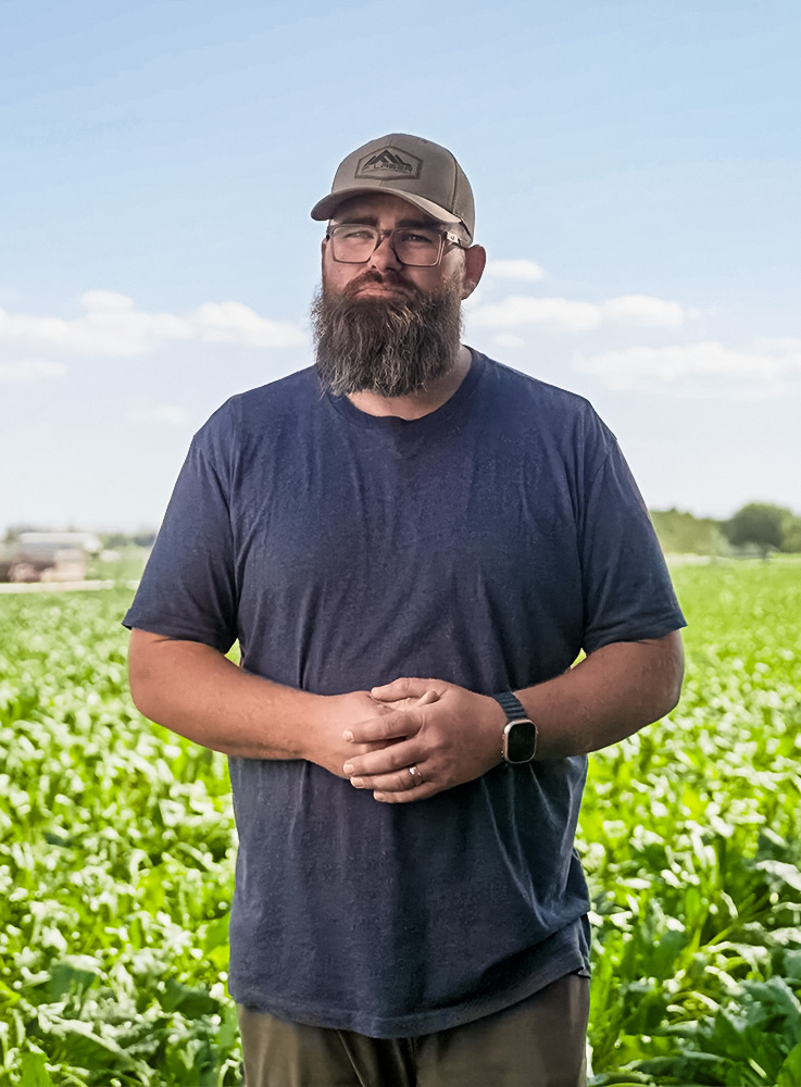 A man with a beard, glasses, and a tan cap stands in front of a green field under a blue sky. He is wearing a navy t-shirt, a smartwatch, and has his hands folded in front of him.