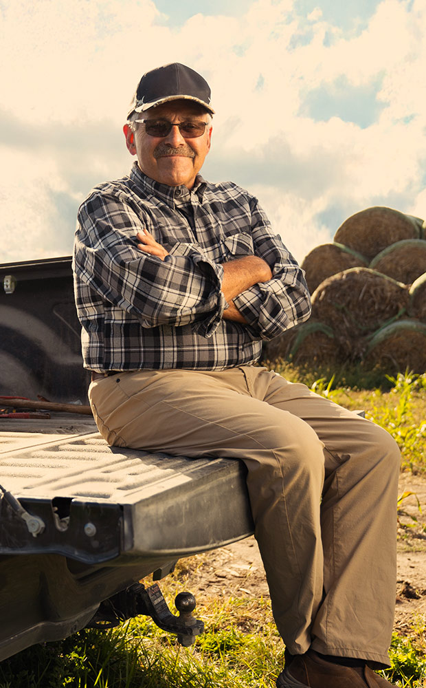 An older man wearing glasses, a black cap, and plaid shirt sits with arms crossed on the back of a pickup truck in a rural field, with hay bales and cloudy sky in the background.