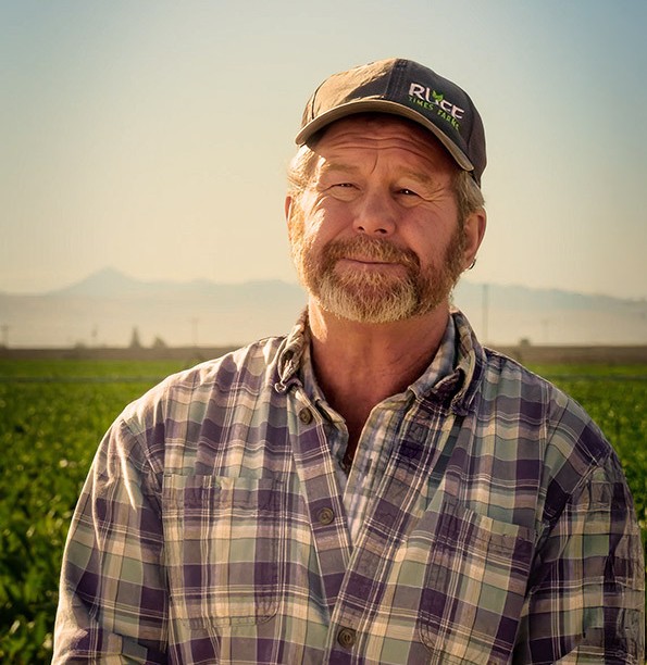 A middle-aged man with a beard wears a plaid shirt and a cap that says RICE. He stands outdoors in front of a green field, where glyphosate weed control helps keep crops healthy, with mountains and a clear sky in the background.
