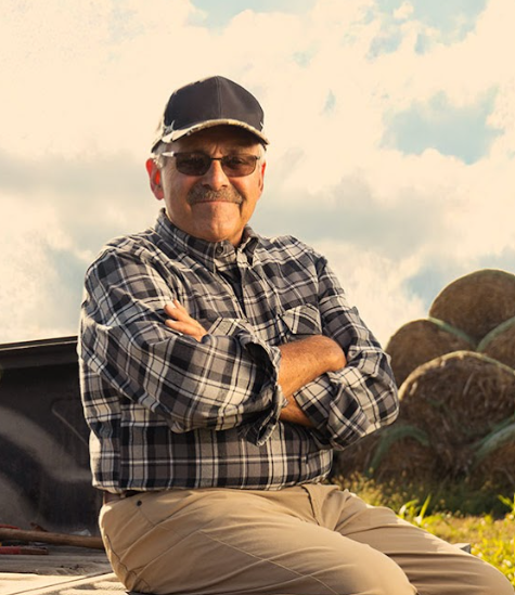 An older man wearing a plaid shirt, sunglasses, and a cap sits outdoors with his arms crossed, smiling. Hay bales and a partly cloudy sky reflect the evolving landscape of modern agriculture in the background.