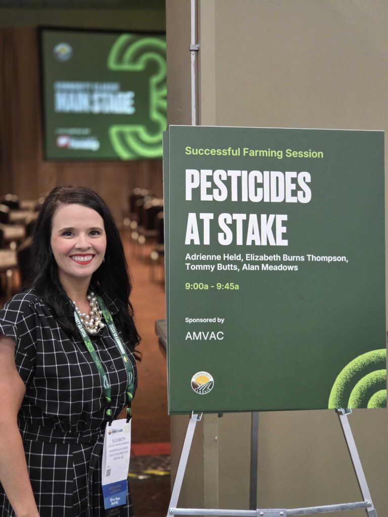 A woman with long dark hair, wearing a black and white checkered dress, stands smiling next to a green sign for a Pesticides at Stake farming session at a conference. Tables and chairs are visible in the background.