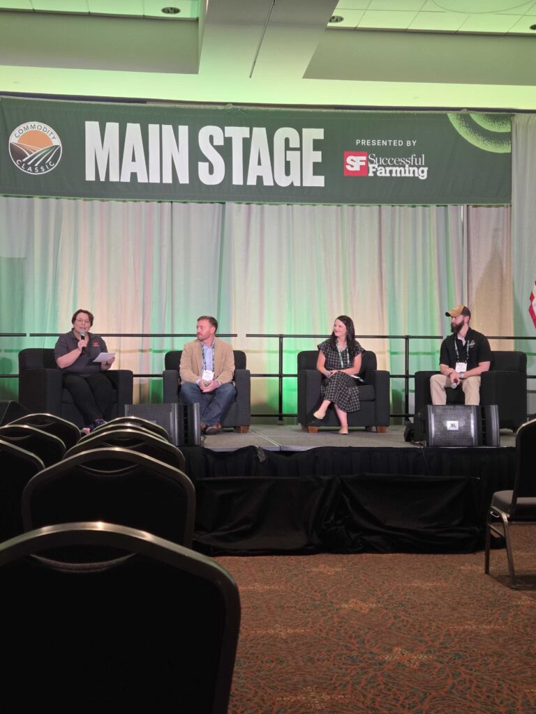 Four people sit on stage chairs under a Main Stage sign at an event, speaking in a panel discussion. The backdrop includes curtains and branding for Successful Farming. Empty audience chairs are visible in the foreground.