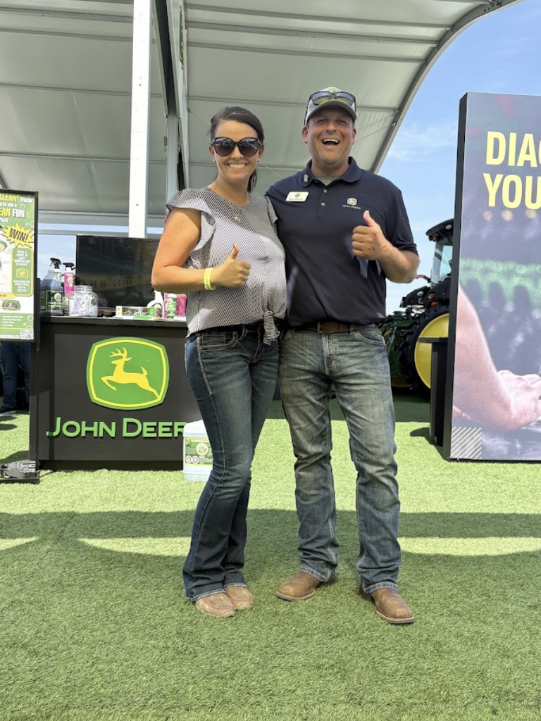 Two people stand side by side on artificial grass, smiling and giving thumbs up. They are in front of a John Deere display booth at an outdoor event. Both are wearing sunglasses and casual clothes.