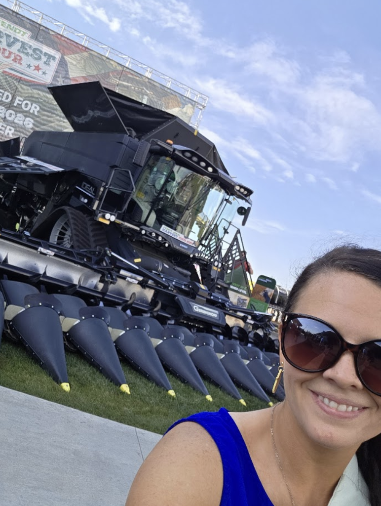 A woman in sunglasses and a blue top smiles for a selfie in front of a large black combine harvester at an outdoor event on a sunny day.