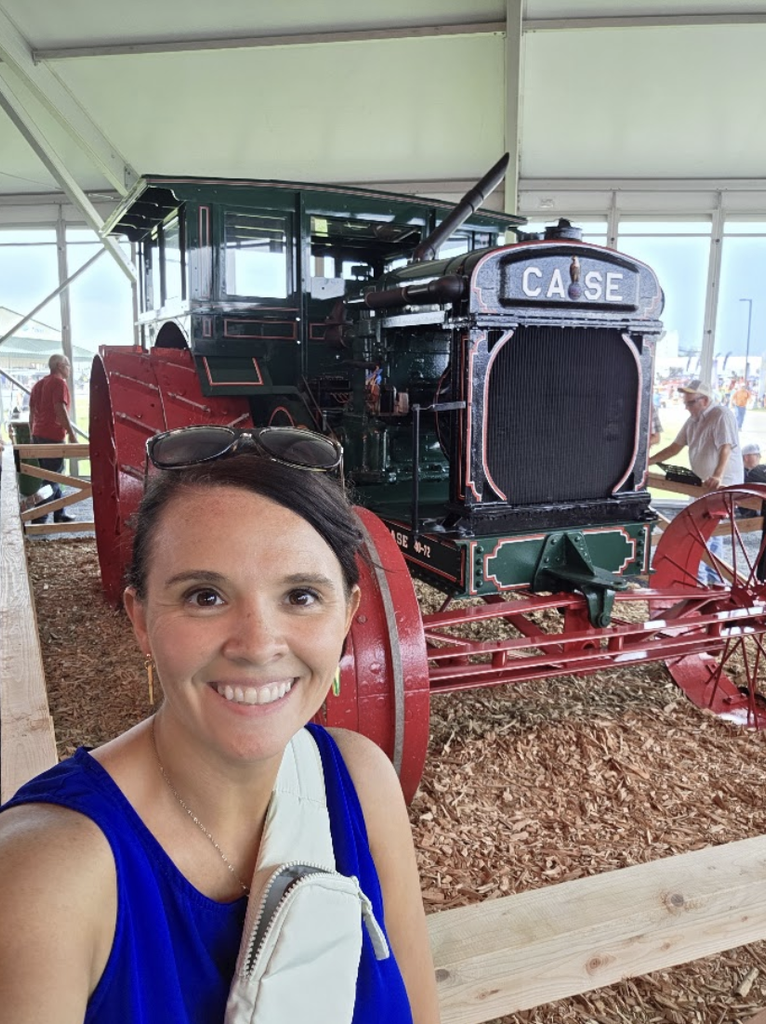 A woman in a blue top smiles in front of a vintage green and red Case tractor displayed indoors on wood chips.