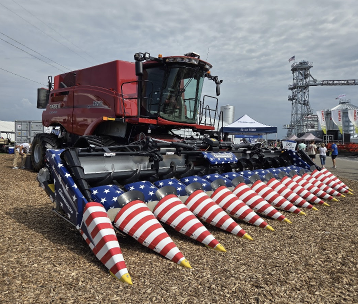 A red combine harvester with a header decorated in a USA stars-and-stripes pattern is displayed at an outdoor event with wood chip ground and a few tents and structures in the background.